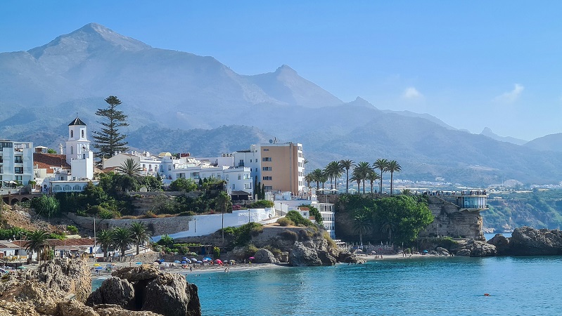 Ljuvliga Nerja Vitkalkade hus vid klippor i Nerja. Turkost hav och mäktiga berg i bakgrunden.