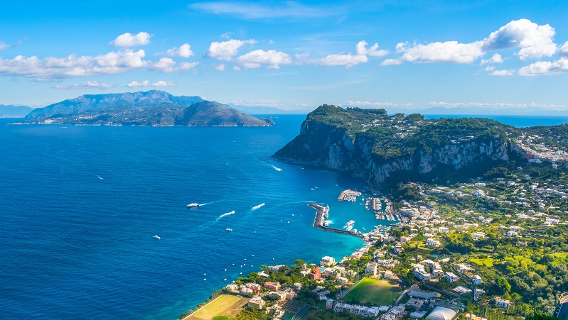 Vy över Capri från Villa San Michele Flygfoto över Capri med klippor, hav, hamn och bebyggelse under en klarblå himmel med vita moln.