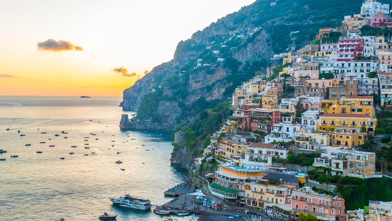 Sorrento Färgglada hus på brant klippa vid havet i solnedgång, med båtar i vattnet nedanför.