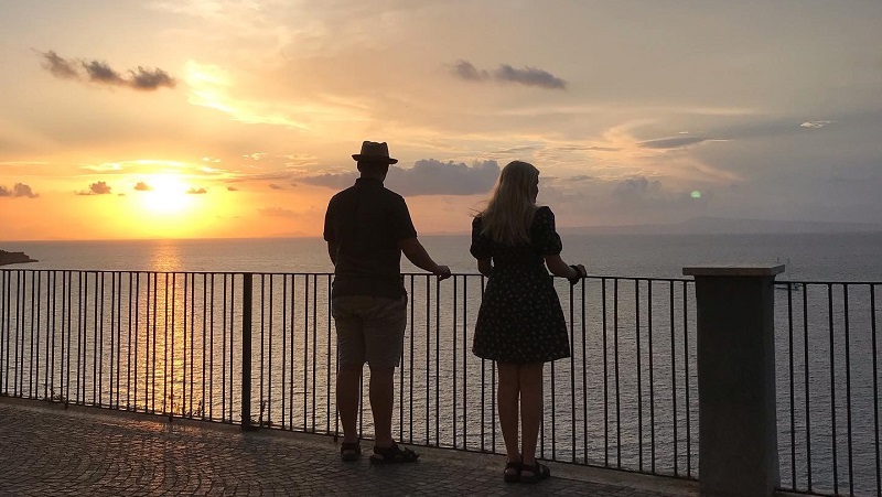 Utsikt i Sorrento Två personer ser ut över havet från en balkong i solnedgången, med gyllene himmel och lugnt vatten.