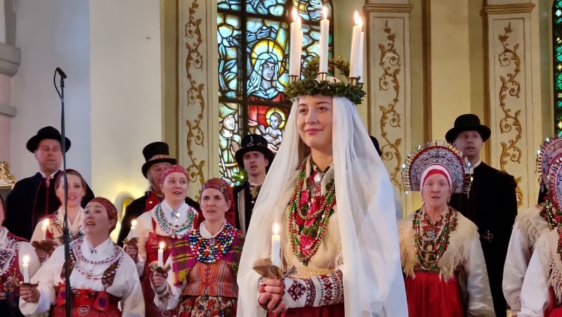 Konserten Dalarnas Lussibrud Människor som är klädda i röda och vita folkdräkter. Kvinna med ljuskrona och vit slöja framför dem.