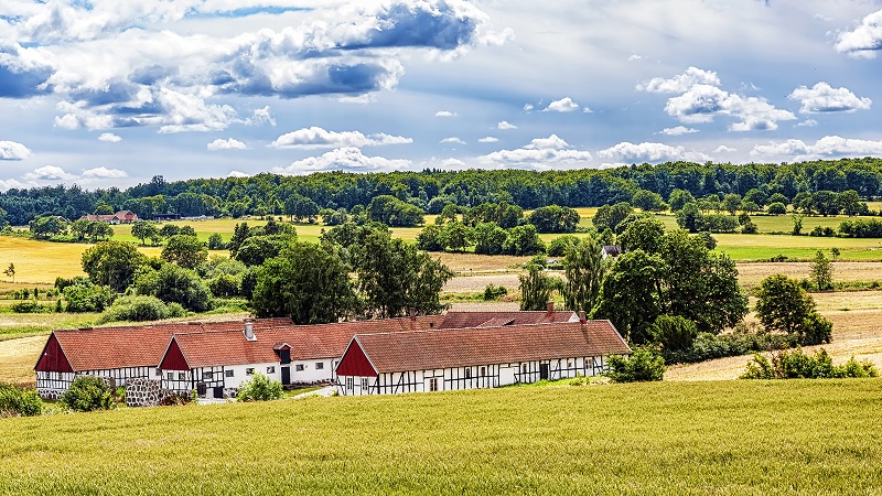 Lång vit korsvirkesgård med röda tak i böljande skånskt landskap under dramatisk himmel med moln.
