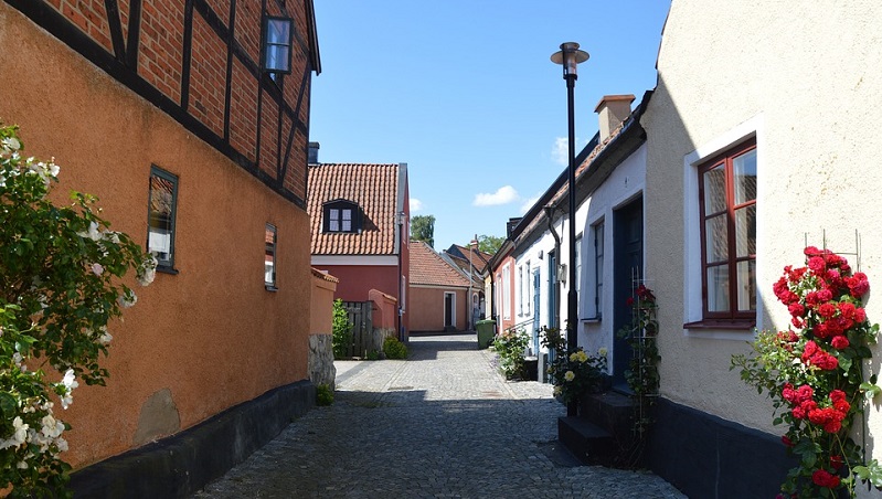 Simrishamn Charmig gränd i Simrishamn med färgglada hus, kullerstensgata, röda rosor och blå himmel.