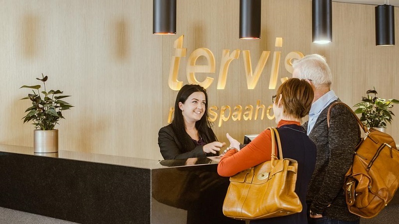 Leende receptionist välkomnar två gäster vid disken på Tervis spa med varm och modern inredning i bakgrunden.