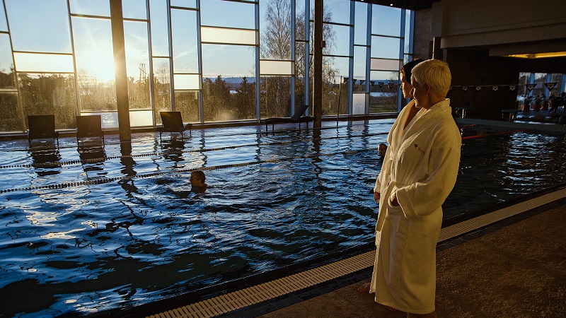 Person i morgonrock vid inomhuspool i solnedgång, med simmande individ och stora fönster som släpper in ljus.