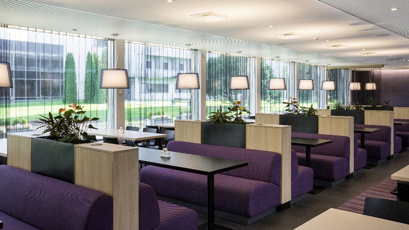 Modern restaurang med fönstervägg, lila soffgrupper, bordslampor och grön utsikt utanför.