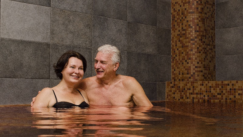 Leende par i inomhuspool med mörk kakelvägg, avslappnad atmosfär och närhet i fokus.