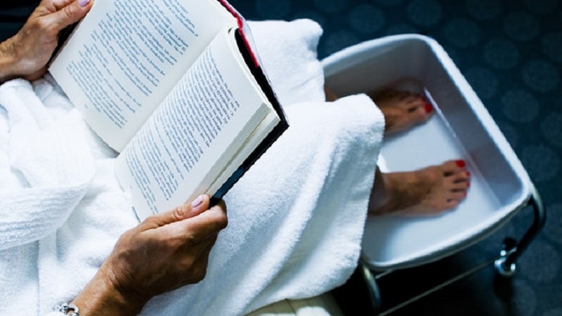 Person i vit morgonrock läser bok meden fötterna vilar i fotbad.