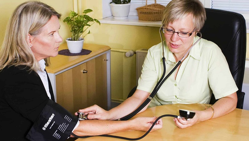 Sjukvårdspersonal mäter blodtryck på patient i lugn, hemtrevlig miljö med växt och träinredning i bakgrunden.