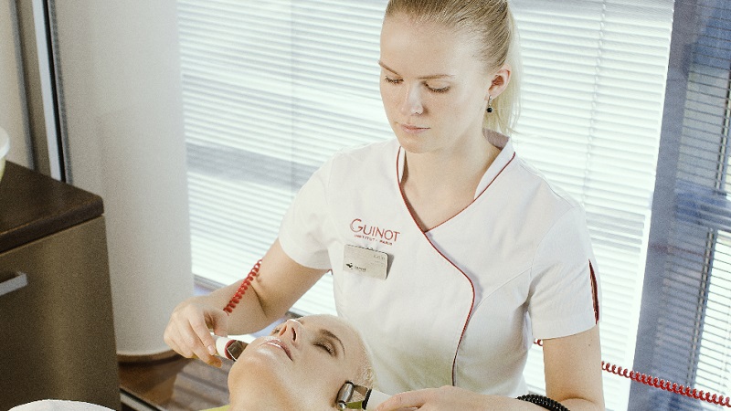 Hudterapeut utför ansiktsbehandling med apparatur i ljus behandlingsmiljö, kunden ligger avslappnad och blundar.