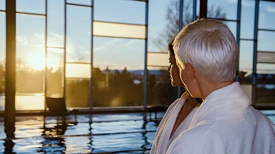 Två personer i vita morgonrockar vid poolkant, blickar ut genom stora fönster mot solnedgången utanför.