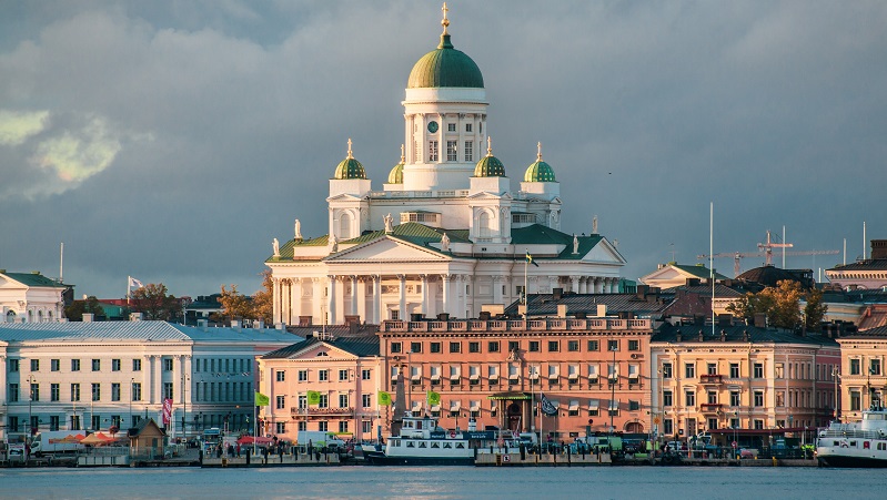 Vit domkyrka i Helsingfors, Finland, lyser upp bakom färgglada byggnader vid vattnet under en blågrå himmel.