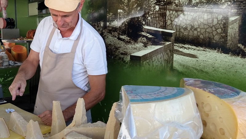 Marknaden i San Casciano Man i vit keps och förkläde skär upp ostbitar vid ett bord fyllt med stora ostar i solskenet.