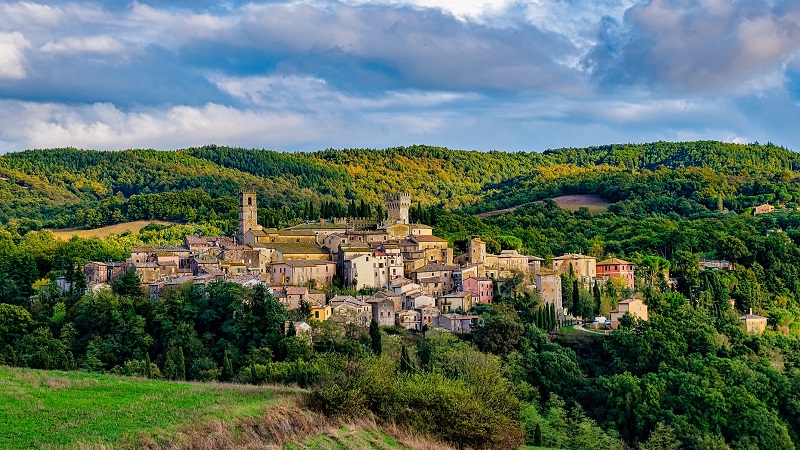 Bild: San Casciano Färgglad by på en kulle omgiven av grönskande skog och böljande landskap under dramatisk himmel.