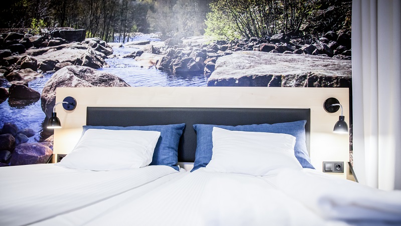 En bäddad hotellsäng med blå kuddar, naturmotiv med forsande vatten som fond bakom sänggaveln.