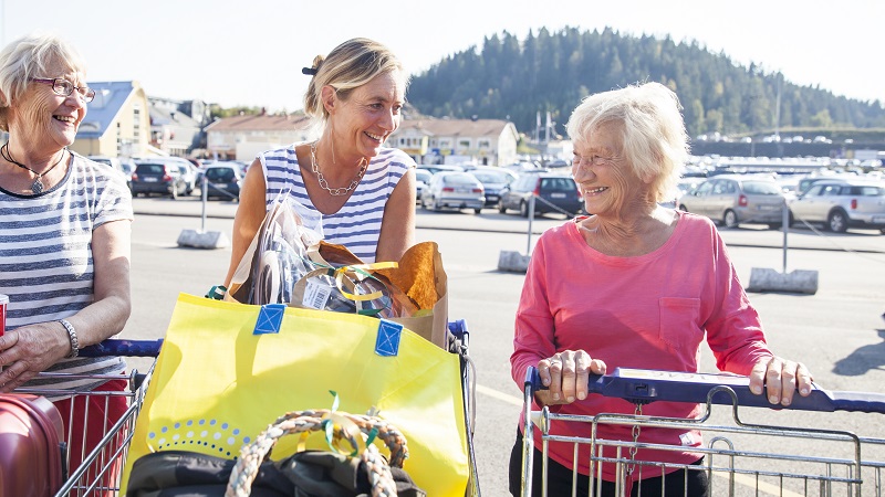 Tre glada kvinnor med kundvagnar fulla med varor, skrattar tillsammans utanför ett shoppingområde.