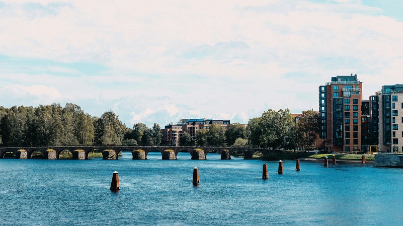 Karlstad Stenbro över klarblått vatten i Karlstad. I bakgrunden syns moderna bostadshus och grönska under en ljus molnig himmel.
