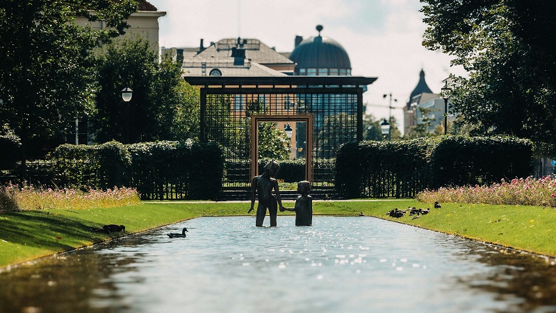 Karlstad Skulptur av 2 personer hand i hand i en vattenspegel. Grönskande parkmiljö med träd, gräs och en smidesgrind i bakgrunden. Stadens byggnader skymtar bakom träden.