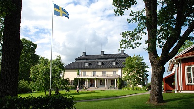 Mårbacka. Vackert stenhus med flaggstång och svensk flagga utanför och gröna gräsmattor.